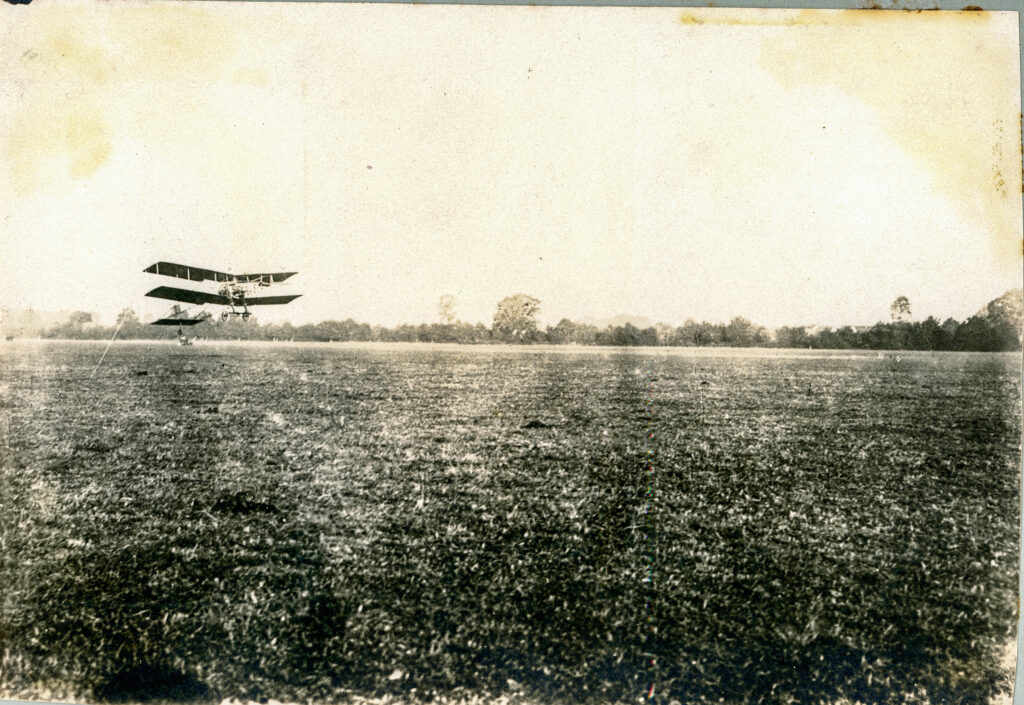 A biplan aeroplane is seen taking flight. The picture is yellowed by time.